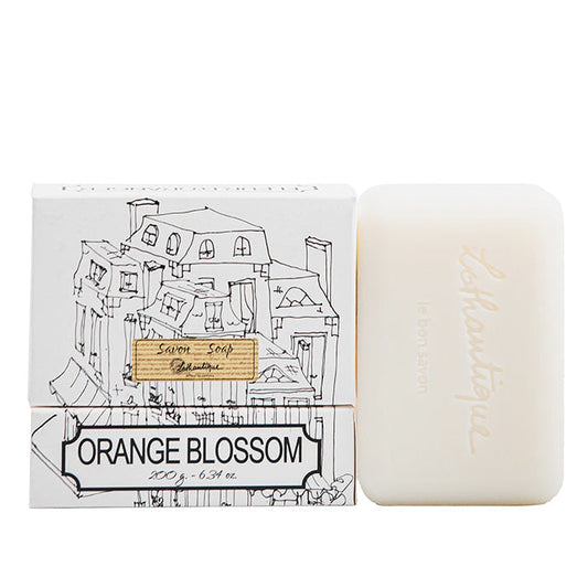 A packaged bar of Authentique Orange Blossom soap with a drawing of a village on the packaging, accompanied by the unwrapped bar displaying the soap's true color.