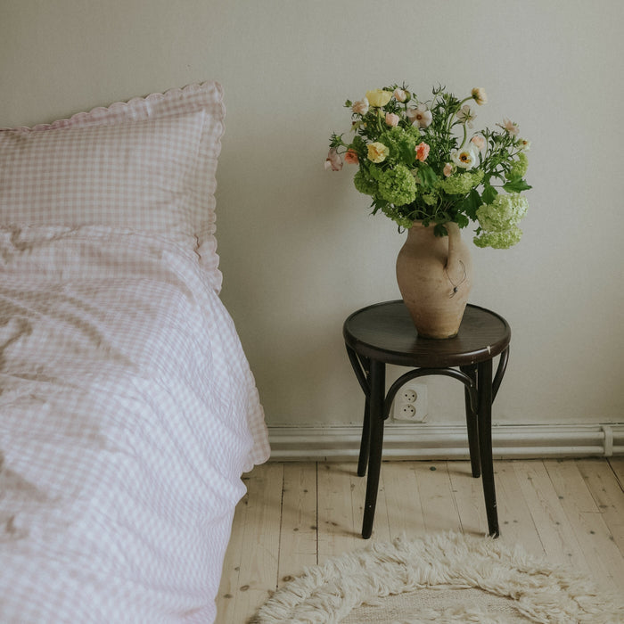 Full shot of a bed with pink gingham print with a side of flowers on a stool