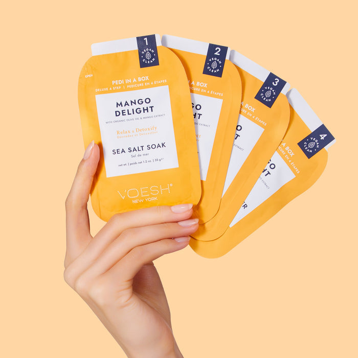 Hand holding Voesh pedicure boxes labeled 'Mango Delight' on a peach background