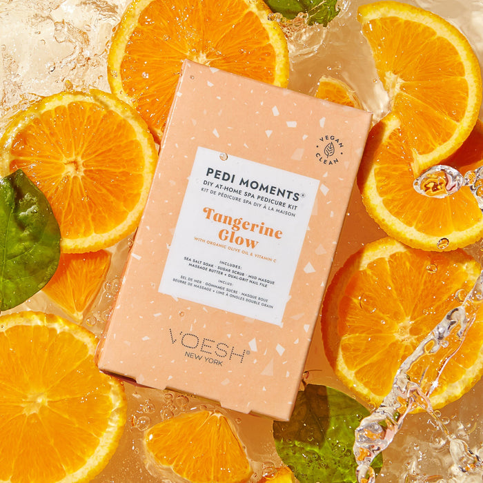 VOESH Pedi Moments Tangerine Glow pedicure kit surrounded by sliced oranges and leaves on a wet surface.